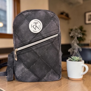Puede incluir: Una bandolera gris estilo denim con cremallera blanca y un parche circular blanco con un logotipo. La bolsa tiene un patrón acolchado y una correa pequeña. Una taza blanca con una planta suculenta está sobre una mesa de madera.