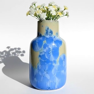 May include: A ceramic vase with a blue and cream glaze, filled with small white and yellow flowers. The vase has a rounded body and a narrow neck. The blue glaze features a crystalline pattern. The vase is approximately 8 inches tall.