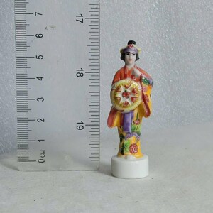 May include: A small, hand-painted porcelain figurine of a person in traditional Asian attire. The figure is approximately 3 inches tall and holds a decorative wheel. The figurine features a yellow, red, and purple color scheme with floral accents.