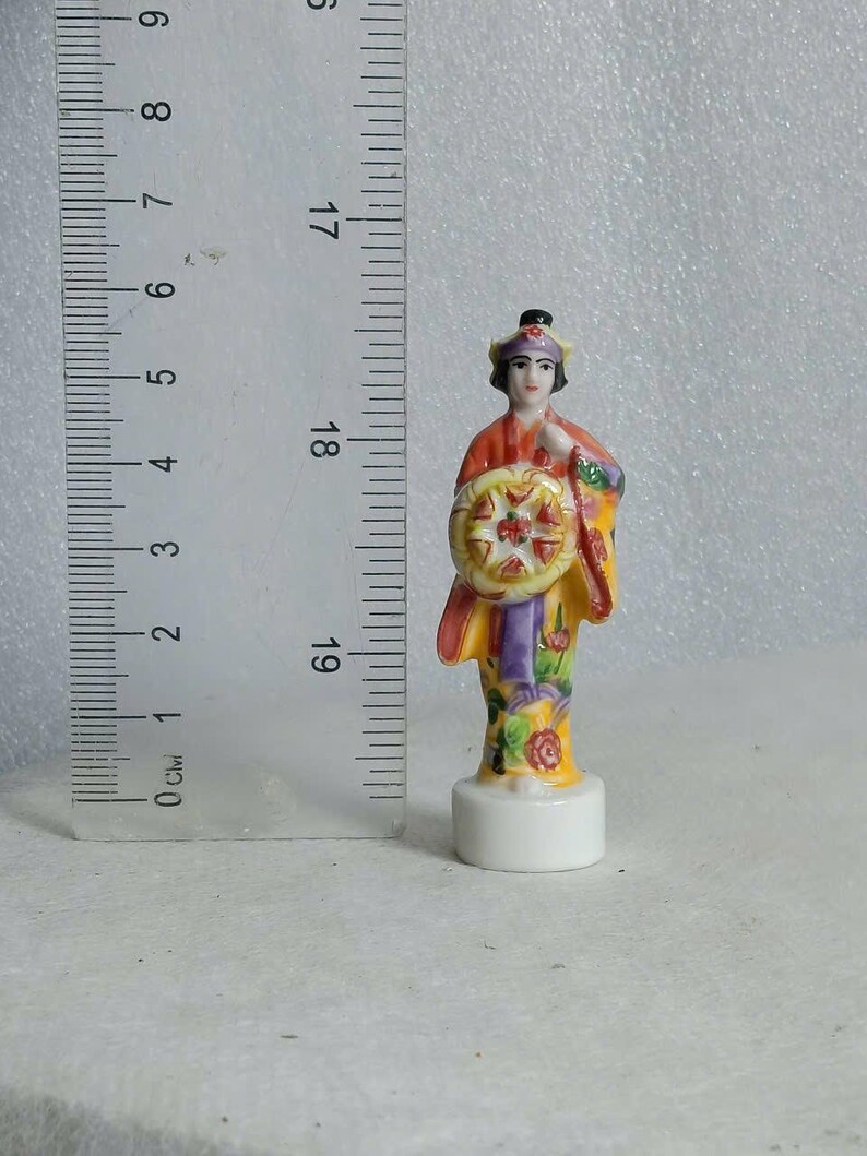May include: A small, hand-painted porcelain figurine of a person in traditional Asian attire. The figure is approximately 3 inches tall and holds a decorative wheel. The figurine features a yellow, red, and purple color scheme with floral accents.