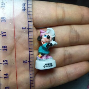 May include: A small Minnie Mouse figurine, approximately 4.4 cm tall, in a teal dress, pink shoes, and a white and pink bow. The figurine is on a white base with the text "MINNIE &copy; DISNEY".