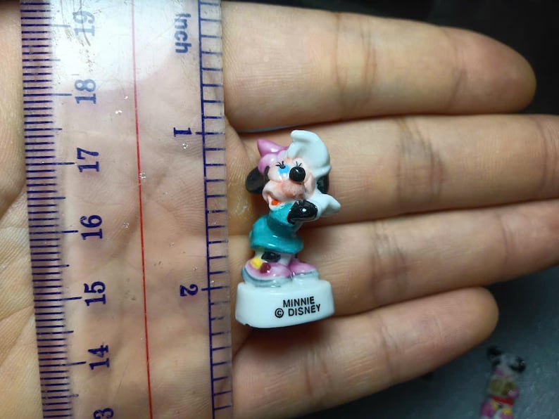 May include: A small Minnie Mouse figurine, approximately 4.4 cm tall, in a teal dress, pink shoes, and a white and pink bow. The figurine is on a white base with the text "MINNIE &copy; DISNEY".