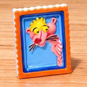 May include: A ceramic Pink Panther figurine in a rectangular frame. The frame has an orange scalloped edge, a blue inner border, and a blue background. The Pink Panther is pink with a yellow head and black whiskers.