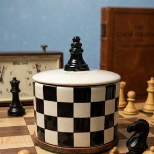 Puede incluir: Un tarro de cerámica con un patrón de tablero de ajedrez en blanco y negro, coronado con una pieza de ajedrez negra. El tarro está sobre una superficie de madera con piezas de ajedrez, un reloj y un libro al fondo. El tarro mide aproximadamente 13 cm de alto.