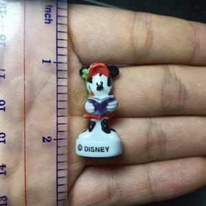 May include: A small Disney Minnie Mouse figurine, approximately 4.4 cm tall, holding a book. Minnie is wearing a red and white outfit with a red and green hat. The base is white with the "&copy; DISNEY" logo.