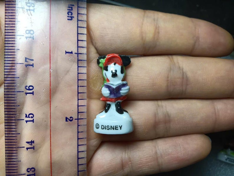 May include: A small Disney Minnie Mouse figurine, approximately 4.4 cm tall, holding a book. Minnie is wearing a red and white outfit with a red and green hat. The base is white with the "&copy; DISNEY" logo.