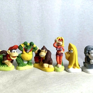 May include: A set of colourful figurines depicting cartoon characters. The collection includes a monkey in a red vest, a green crocodile in a yellow shirt, and a banana-shaped figure. The figures are on small, green and yellow bases.