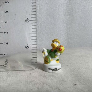 May include: A small Donald Duck figurine, wearing a green jacket and yellow hat, holding a bouquet of flowers. The figurine is on a white base with text. A ruler is next to the figurine, showing centimeters.