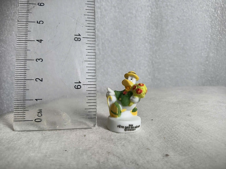 May include: A small Donald Duck figurine, wearing a green jacket and yellow hat, holding a bouquet of flowers. The figurine is on a white base with text. A ruler is next to the figurine, showing centimeters.