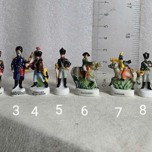 Może przedstawiać: Kolekcja dziesięciu miniaturowych figurek porcelanowych przedstawiających żołnierzy w historycznych mundurach. Figurki są pomalowane szczegółowymi kolorami i stoją na małych białych podstawach. Za figurkami umieszczono linijkę.