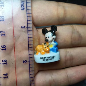 May include: A small ceramic Baby Mickey Mouse figurine, approximately 2.5 cm tall, with a brown and orange animal figure. Mickey is wearing blue and white clothing. The base is white with the text "BABY MICKEY &copy; DISNEY".