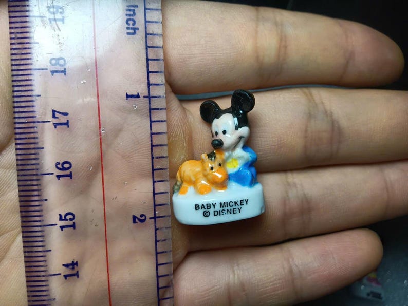 May include: A small ceramic Baby Mickey Mouse figurine, approximately 2.5 cm tall, with a brown and orange animal figure. Mickey is wearing blue and white clothing. The base is white with the text "BABY MICKEY &copy; DISNEY".