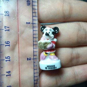 May include: A small Minnie Mouse figurine, approximately 5 cm tall, is held next to a ruler. The figurine is wearing a red and pink outfit and holding a basket. The base of the figurine reads "MINNIE DISNEY".