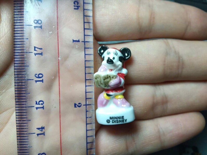 May include: A small Minnie Mouse figurine, approximately 5 cm tall, is held next to a ruler. The figurine is wearing a red and pink outfit and holding a basket. The base of the figurine reads "MINNIE DISNEY".
