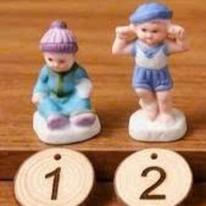 May include: Two small porcelain figurines of children, one seated and wearing a blue and teal outfit with a purple hat, the other standing and covering their ears. Two wooden discs with the numbers 1 and 2 are in front.
