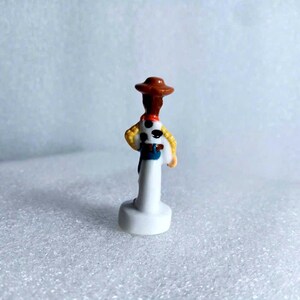 Pu&ograve; includere: Piccola e dettagliata statuina del personaggio Woody del film d'animazione Toy Story. La statuina presenta un cappello da cowboy marrone, una camicia gialla, un fazzoletto rosso e un gilet blu. La figura &egrave; montata su una base bianca.