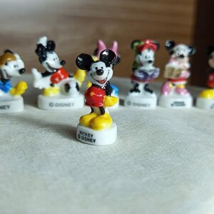 Puede incluir: Una colecci&oacute;n de peque&ntilde;as figuras de Mickey Mouse. La figura central muestra a Mickey Mouse con pantalones cortos rojos, zapatos amarillos y una chaqueta negra. Cada figura est&aacute; sobre una base blanca con "MICKEY &copy; DISNEY".