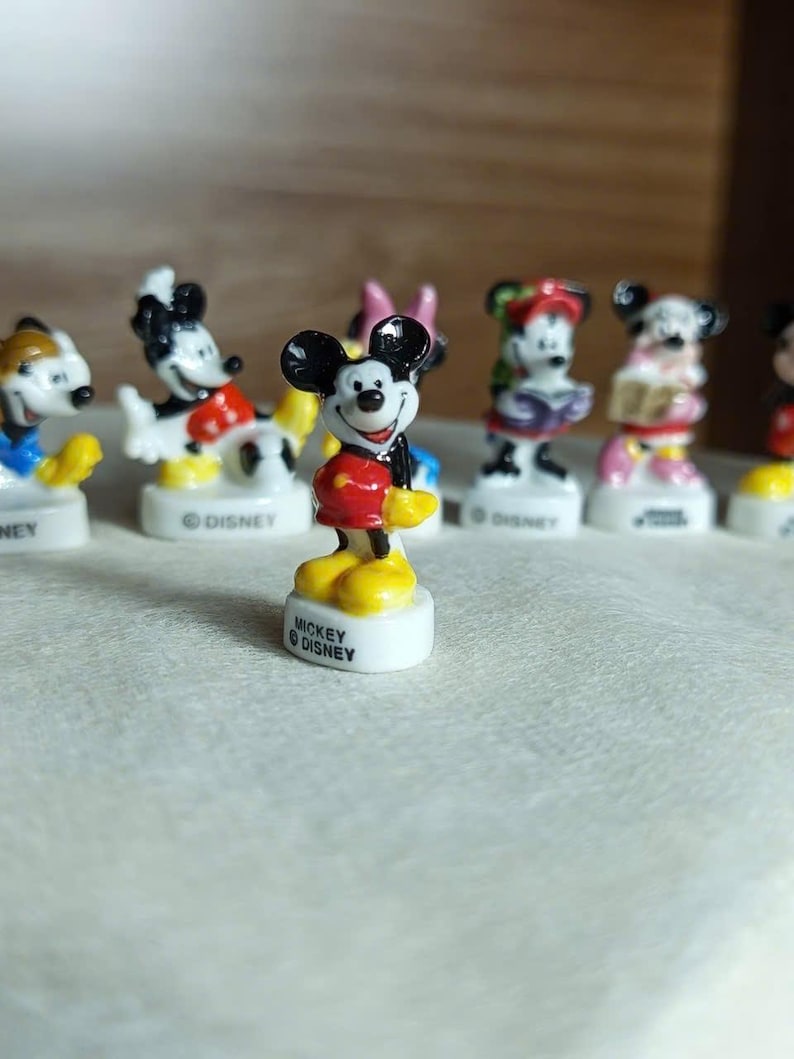 Puede incluir: Una colecci&oacute;n de peque&ntilde;as figuras de Mickey Mouse. La figura central muestra a Mickey Mouse con pantalones cortos rojos, zapatos amarillos y una chaqueta negra. Cada figura est&aacute; sobre una base blanca con "MICKEY &copy; DISNEY".