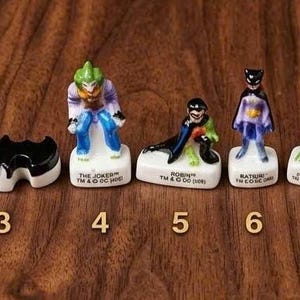 May include: A collection of nine ceramic Batman character figurines, including Catwoman, Batman, The Joker, and The Penguin, on a wooden surface. Each figurine is detailed with vibrant colors and stands on a white base.