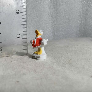 May include: A small, white and yellow Donald Duck figurine wearing a red coat. The figurine is approximately 2 inches tall and stands on a white base. The figurine is next to a ruler.
