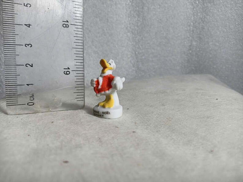 May include: A small, white and yellow Donald Duck figurine wearing a red coat. The figurine is approximately 2 inches tall and stands on a white base. The figurine is next to a ruler.