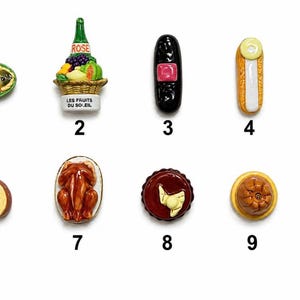 May include: A collection of ten ceramic food-themed miniatures. Includes a fish dish, a bottle with fruit, a black object with a pink square, a hot dog, a nut, a pastry roll, a roasted chicken, a tart, a pie, and a plate of cookies.