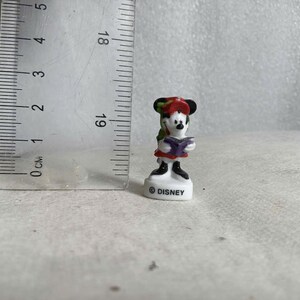 May include: A small Minnie Mouse figurine, approximately 2 inches tall, is depicted in a reading pose. She wears a red dress, a green and red hat, and holds a purple book. The base is white with the "&copy; DISNEY" logo.