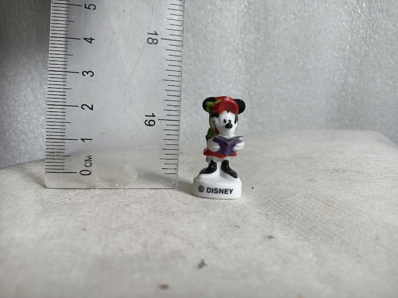 May include: A small Minnie Mouse figurine, approximately 2 inches tall, is depicted in a reading pose. She wears a red dress, a green and red hat, and holds a purple book. The base is white with the "&copy; DISNEY" logo.