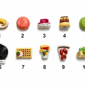 May include: A collection of ten miniature food-themed figurines. The figurines include a croissant, a pink macaron, a small jar, a layered cake, a green macaron, a licorice wheel, a waffle, a dessert, a pizza slice, and a small vehicle.