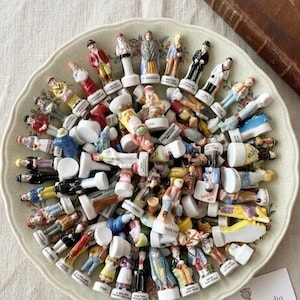Può includere: Una collezione di piccole statuette in ceramica dipinte a mano, o "fèves", in vari abiti e pose, disposte su un piatto decorativo. Le statuette sono prevalentemente bianche, con dettagli colorati. Un libro d'epoca è sullo sfondo.