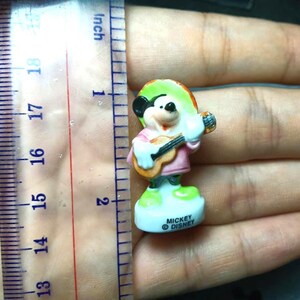 May include: A small Mickey Mouse figurine, approximately 5 cm tall, dressed in a pink shirt and sombrero, playing a guitar. The figurine is on a white base with the text "Mickey Disney".