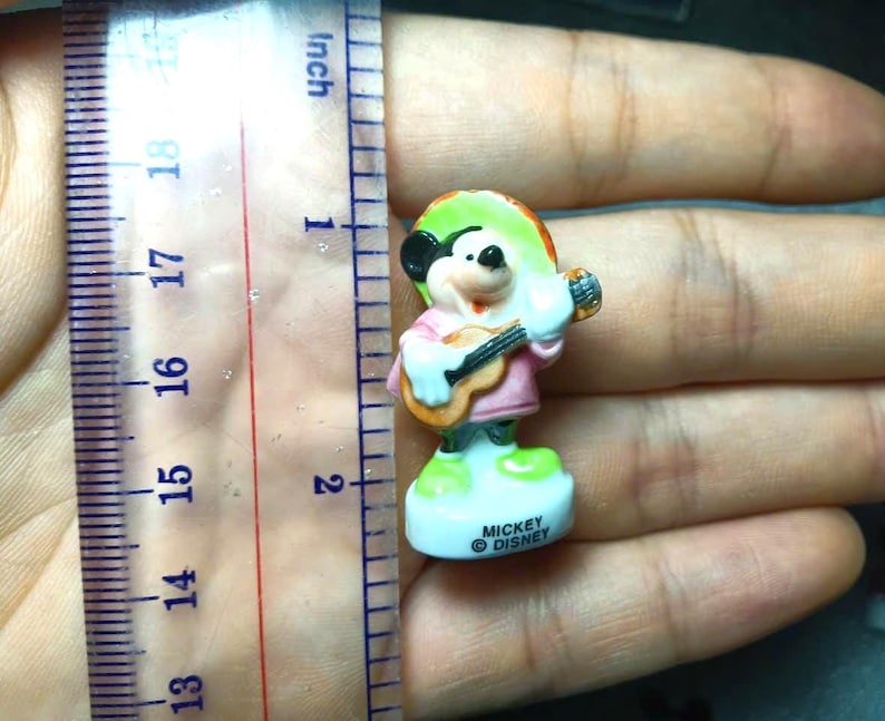 May include: A small Mickey Mouse figurine, approximately 5 cm tall, dressed in a pink shirt and sombrero, playing a guitar. The figurine is on a white base with the text "Mickey Disney".