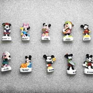 May include: A set of ten miniature Disney character figurines. The figures depict Mickey Mouse, Minnie Mouse, and other characters in different outfits. Each figurine stands on a white base with the Disney name.