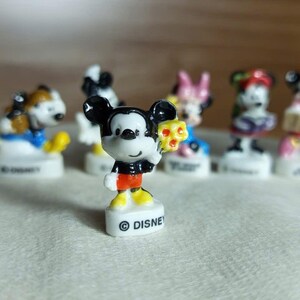 Puede incluir: Una colecci&oacute;n de peque&ntilde;as figuras de personajes de Disney. Mickey Mouse es la figura central, sosteniendo un objeto amarillo. Mickey lleva pantalones cortos negros, zapatos amarillos y una chaqueta negra. Las figuras est&aacute;n sobre bases blancas con la palabra "DISNEY" impresa.