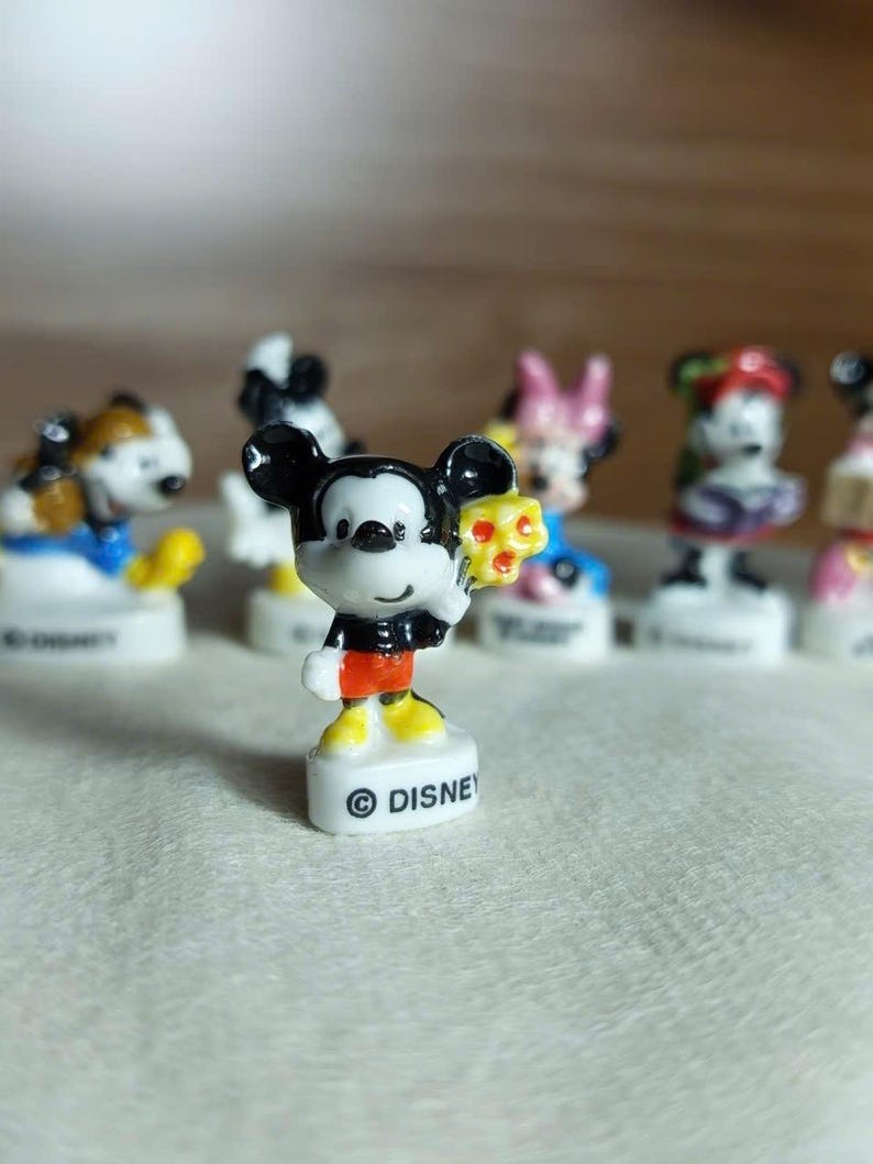 Puede incluir: Una colecci&oacute;n de peque&ntilde;as figuras de personajes de Disney. Mickey Mouse es la figura central, sosteniendo un objeto amarillo. Mickey lleva pantalones cortos negros, zapatos amarillos y una chaqueta negra. Las figuras est&aacute;n sobre bases blancas con la palabra "DISNEY" impresa.