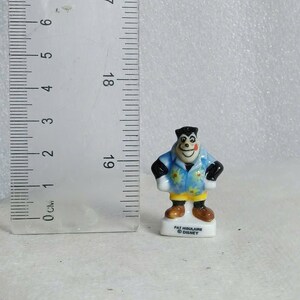 May include: A small, ceramic figurine of a cartoon character, approximately 1.5 inches tall. The figure is wearing a blue shirt with a floral pattern, black sleeves, and brown shoes. The base is white with text that reads "PAT HIGULAIRE &copy; DISNEY".