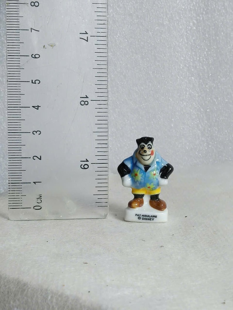 May include: A small, ceramic figurine of a cartoon character, approximately 1.5 inches tall. The figure is wearing a blue shirt with a floral pattern, black sleeves, and brown shoes. The base is white with text that reads "PAT HIGULAIRE &copy; DISNEY".