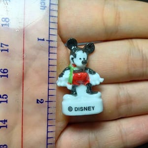 May include: A small Mickey Mouse figurine, approximately 4.4 cm tall, with black ears, a red shirt, and black shoes. The figurine has a white base with the word "DISNEY" printed on it. A green and red scarf is around the neck.