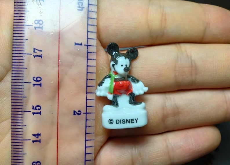May include: A small Mickey Mouse figurine, approximately 4.4 cm tall, with black ears, a red shirt, and black shoes. The figurine has a white base with the word "DISNEY" printed on it. A green and red scarf is around the neck.