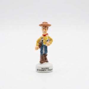 Pu&ograve; includere: Una piccola statuina di Woody di Toy Story della Disney/Pixar. La figura indossa un cappello da cowboy marrone, una camicia a quadri gialla, jeans blu e stivali marroni. La base &egrave; bianca con il nome "WOODY" e il logo Disney/Pixar.