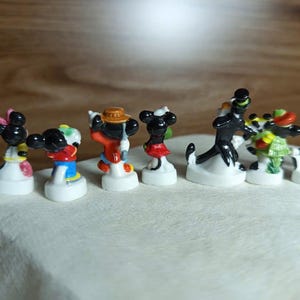 以下が含まれることがあります： 様々なディズニーキャラクターをあしらった、小さくてカラフルなフィギュアのコレクション。フィギュアは白い台座に置かれ、ミッキーマウス、ミニーマウス、その他の有名なキャラクターが含まれています。キャラクターは象徴的な衣装を着ています。