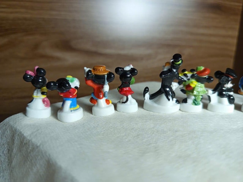 以下が含まれることがあります： 様々なディズニーキャラクターをあしらった、小さくてカラフルなフィギュアのコレクション。フィギュアは白い台座に置かれ、ミッキーマウス、ミニーマウス、その他の有名なキャラクターが含まれています。キャラクターは象徴的な衣装を着ています。