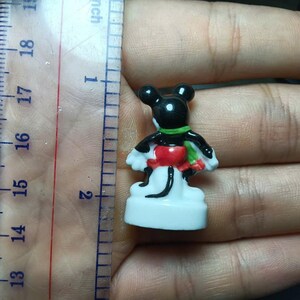 May include: A small, colorful Mickey Mouse figurine, approximately 1.75 inches tall, is held next to a ruler. The figure has a black head and body, white gloves, a red and green outfit, and a white base. The figurine is a collectible item.