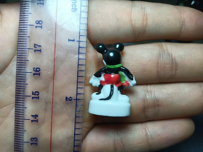 May include: A small, colorful Mickey Mouse figurine, approximately 1.75 inches tall, is held next to a ruler. The figure has a black head and body, white gloves, a red and green outfit, and a white base. The figurine is a collectible item.