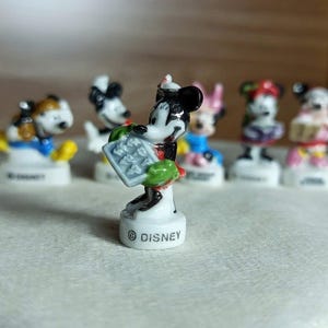 Puede incluir: Colecci&oacute;n de figuras de Disney, con Mickey Mouse y otros personajes. Mickey, en el centro, es blanco y negro, sosteniendo un objeto gris. Las figuras est&aacute;n sobre pedestales blancos, con el logo de Disney. Otros personajes en el fondo.