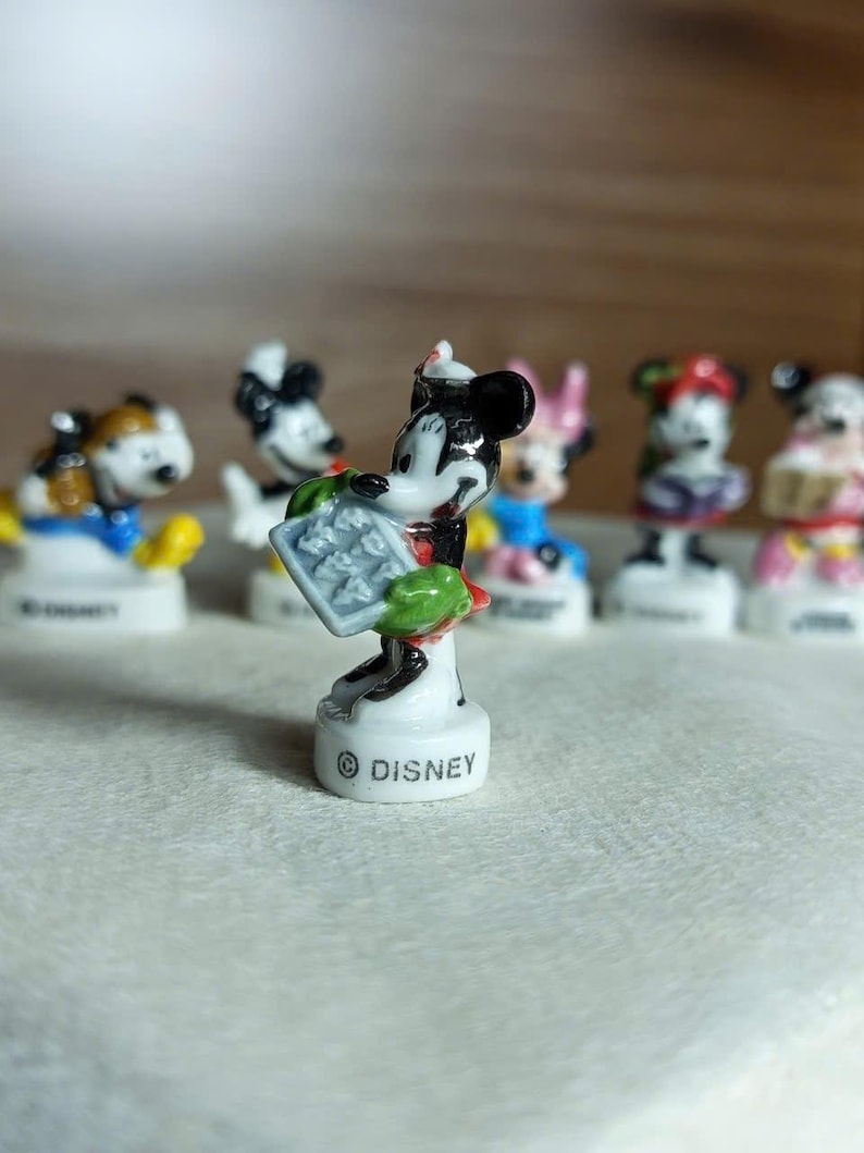 Puede incluir: Colecci&oacute;n de figuras de Disney, con Mickey Mouse y otros personajes. Mickey, en el centro, es blanco y negro, sosteniendo un objeto gris. Las figuras est&aacute;n sobre pedestales blancos, con el logo de Disney. Otros personajes en el fondo.