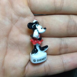 May include: A small, ceramic Minnie Mouse figurine. The figure is black, white, and red, with a white face, black ears, and a red skirt. The base is white with the word "Disney" printed on it.