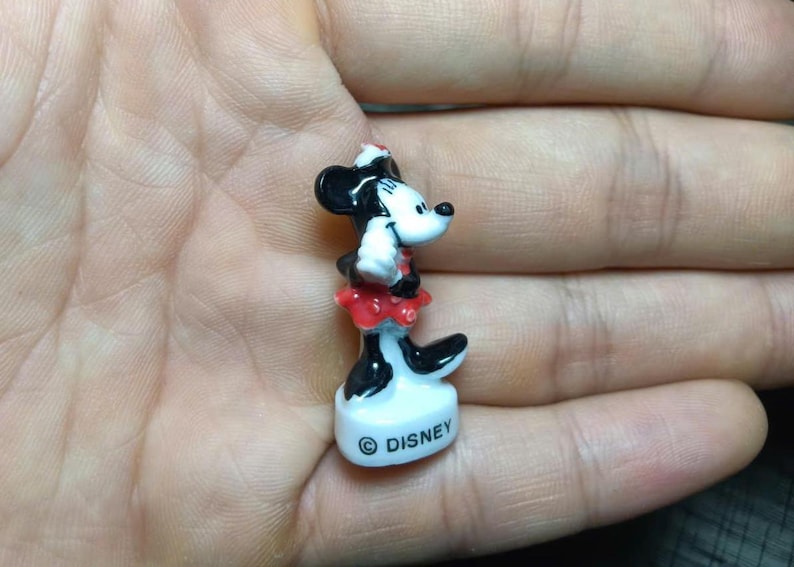 May include: A small, ceramic Minnie Mouse figurine. The figure is black, white, and red, with a white face, black ears, and a red skirt. The base is white with the word "Disney" printed on it.