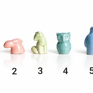 Może przedstawiać: Zestaw sześciu ceramicznych figurek zwierząt w różnych kolorach. Figurki obejmują białego niedźwiedzia, różowego królika, jasnozieloną żabę, turkusową sowę, niebieskiego pingwina i turkusowego jelenia. Każda figurka ma około 5 cm wysokości.