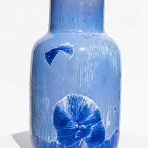 Könnte beinhalten: Eine blaue Keramikvase mit rundem Körper und zylindrischem Hals. Die Vase hat eine strukturierte Oberfläche mit dunkelblauen blütenartigen Mustern und Akzenten. Die Vase wird auf einer weißen Oberfläche präsentiert.